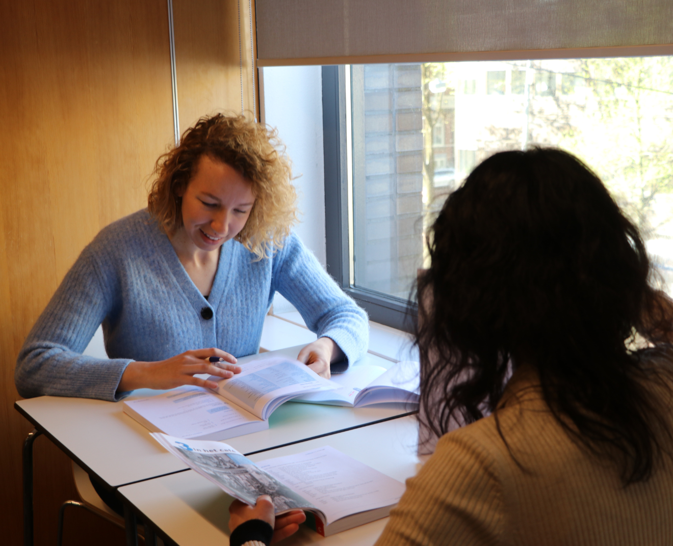 Twee vrouwen zitten aan een tafel bij een raam en werken samen met studieboeken en papieren. De vrouw in de blauwe trui lacht terwijl ze iets aanwijst in haar boek.