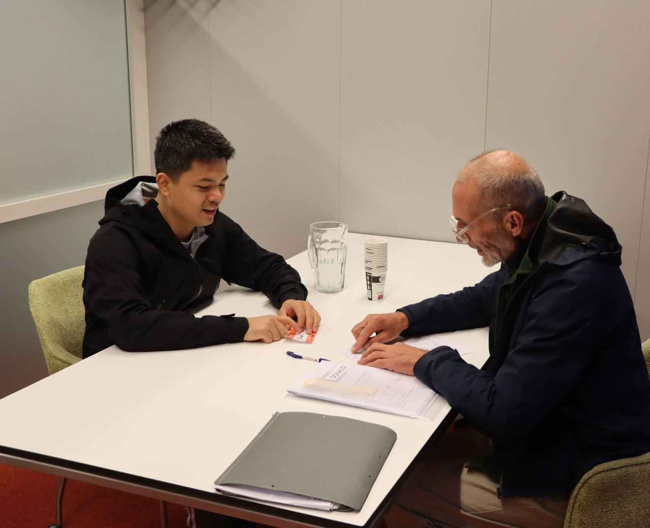 Twee mannen zitten aan een tafel in een gesprek, met papieren en een map voor zich. De jongere man in een zwarte hoodie glimlacht, terwijl de oudere man met bril en baard iets aanwijst op een document.