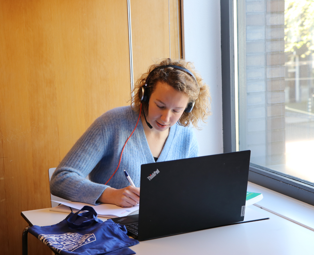 Een vrouw met een krullend blond kapsel en een headset zit aan een tafel bij het raam, werkend op haar laptop en notities makend. Naast haar ligt een blauwe tas en enkele studieboeken.