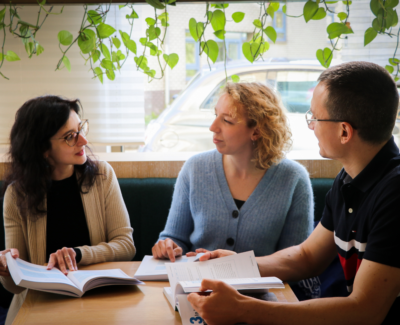 Drie mensen zitten in een caféhoek met groene hangplanten en bespreken hun studieboeken. Ze kijken naar elkaar en lijken in gesprek, terwijl hun boeken open op tafel liggen.