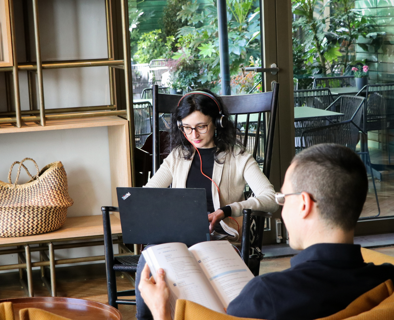 Een vrouw met een headset werkt op haar laptop in een sfeervolle ruimte met veel groen. Voor haar zit een man in een gele fauteuil, lezend in een studieboek.