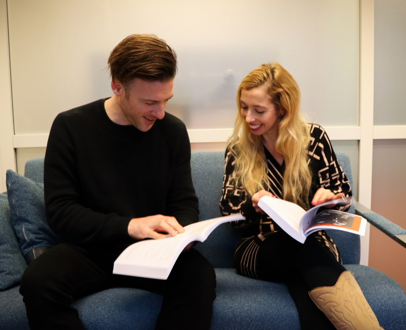 Een man en een vrouw zitten samen op een blauwe bank en lachen terwijl ze in hun studieboeken bladeren. Ze lijken enthousiast iets in de boeken te bespreken.