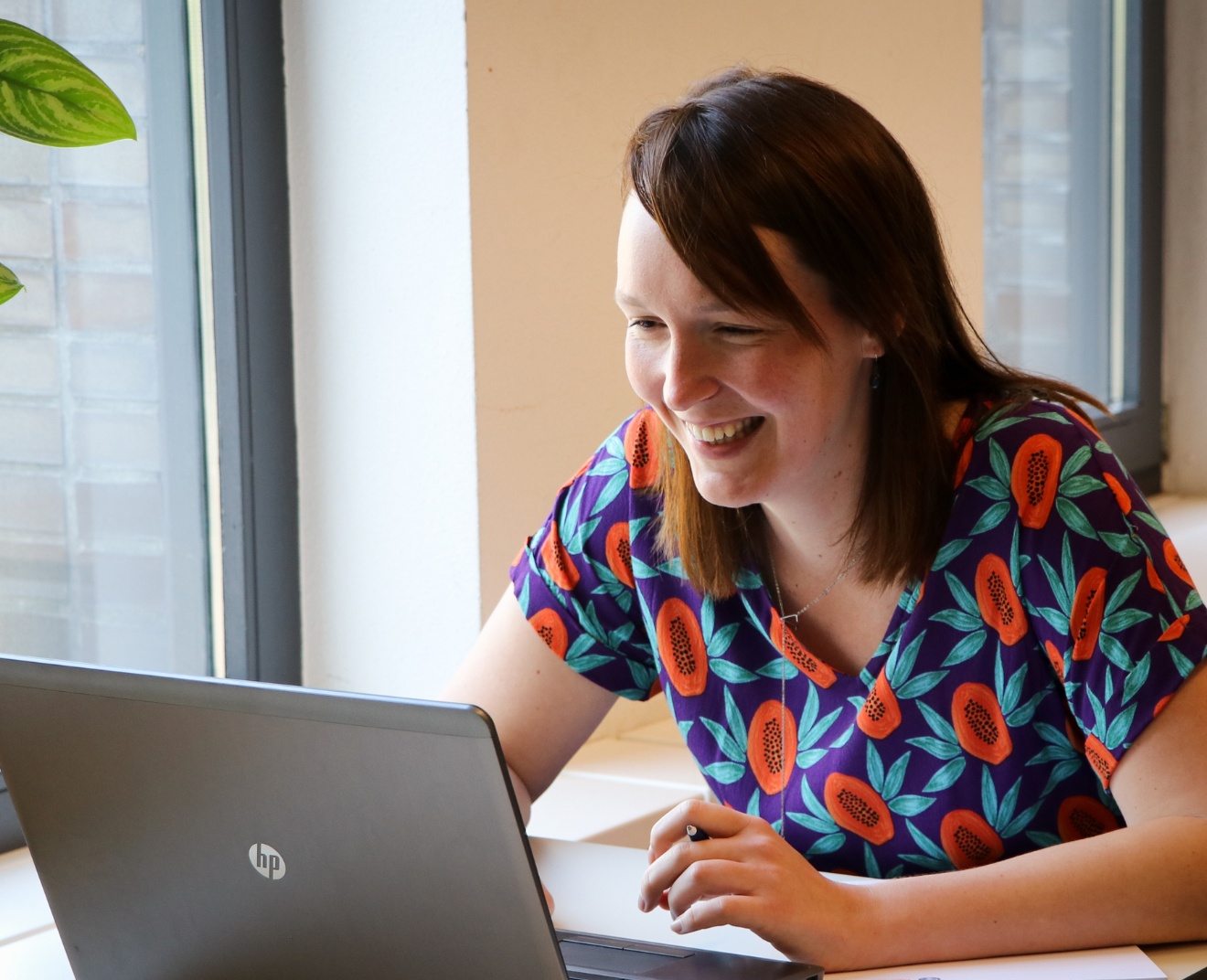 Een lachende vrouw met halflang bruin haar en een kleurrijke blouse met papajaprint werkt op haar laptop. Ze zit bij een raam met natuurlijk licht en een plant op de achtergrond.