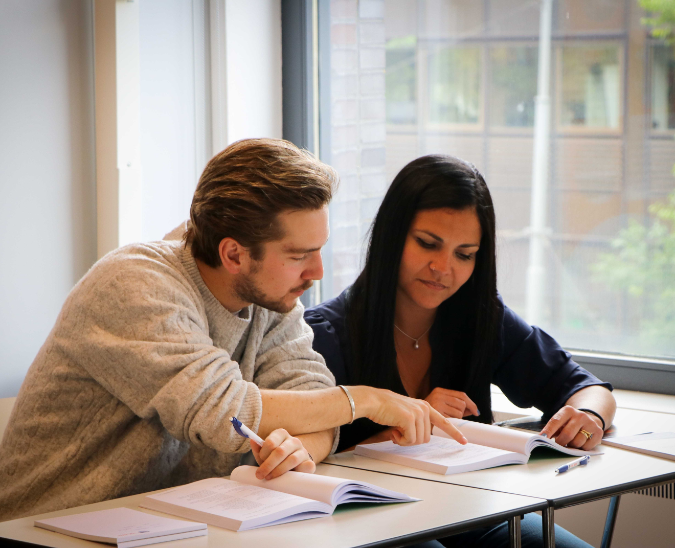 Een man en een vrouw zitten samen aan een tafel en bestuderen een boek. De man wijst iets aan terwijl de vrouw aandachtig meeleest.