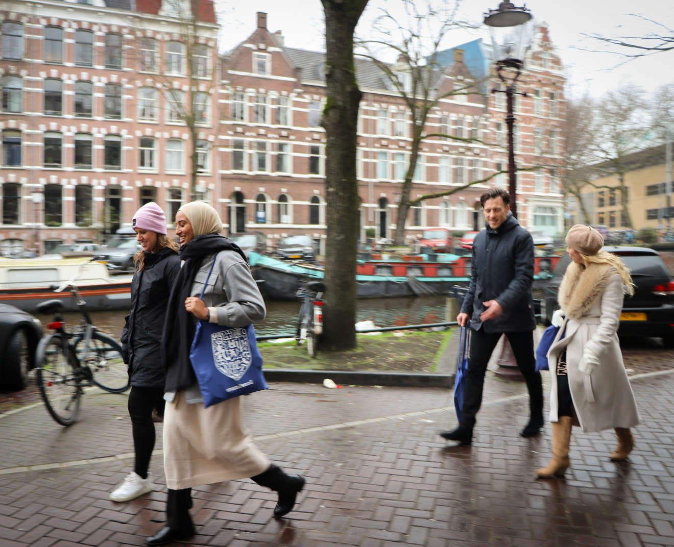 Vier mensen lopen langs een gracht met historische gebouwen en boten. Ze dragen winterkleding en blauwe tassen terwijl ze in gesprek zijn.