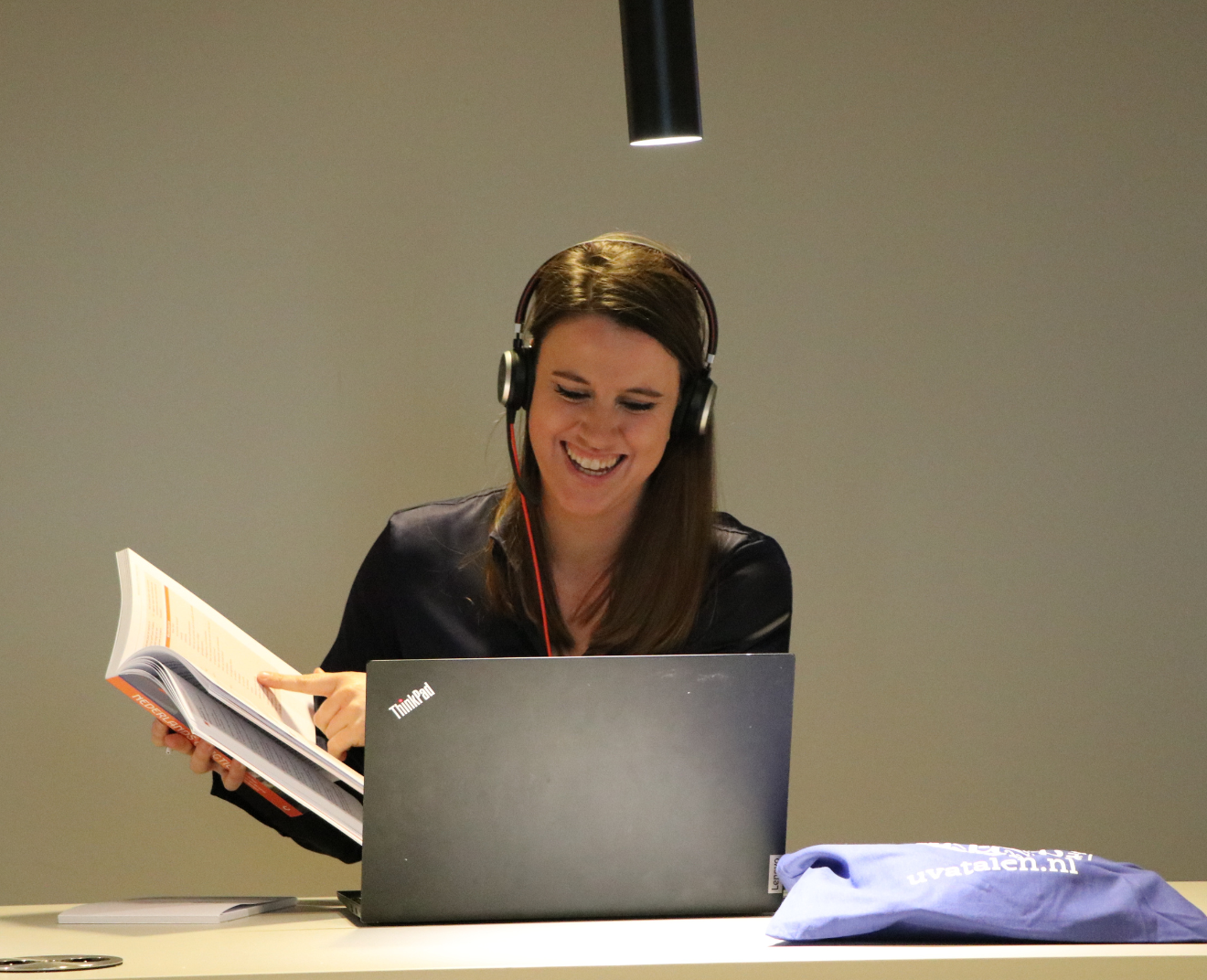 Een lachende vrouw met een headset zit achter een laptop en bladert door een studieboek. Op tafel ligt een blauwe tas met een logo.