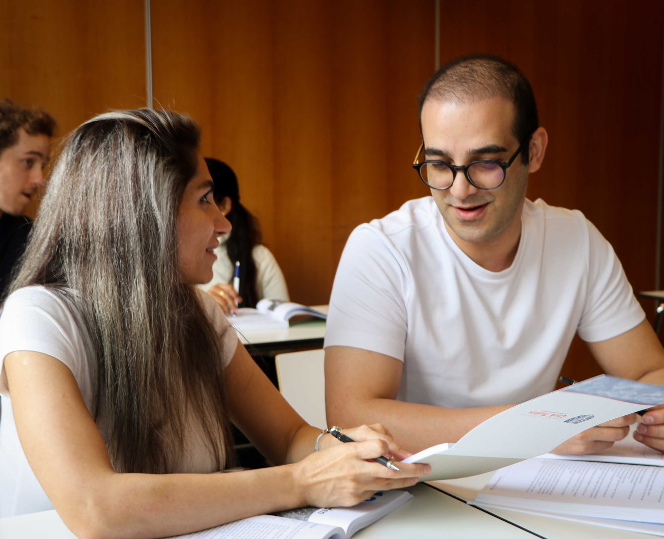 Een man en een vrouw zitten in een klaslokaal en bespreken samen een document. Op de achtergrond werken andere studenten met boeken en notities.