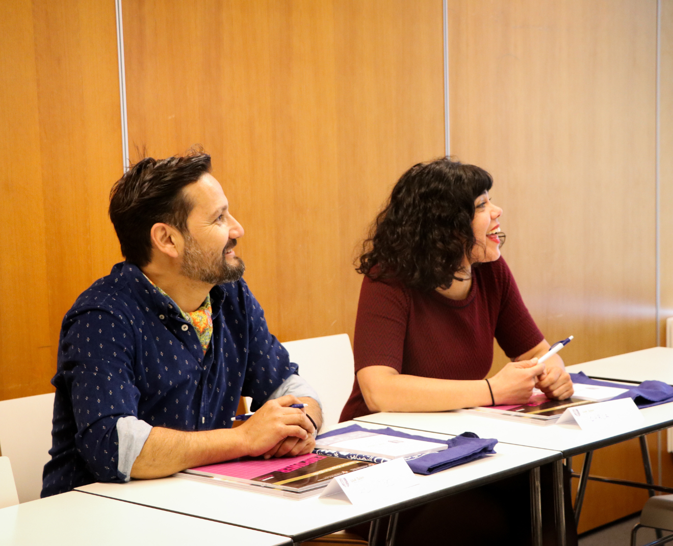 Een man en een vrouw zitten in een klaslokaal en lachen terwijl ze luisteren. Voor hen liggen notities, pennen en tassen op de tafel.
