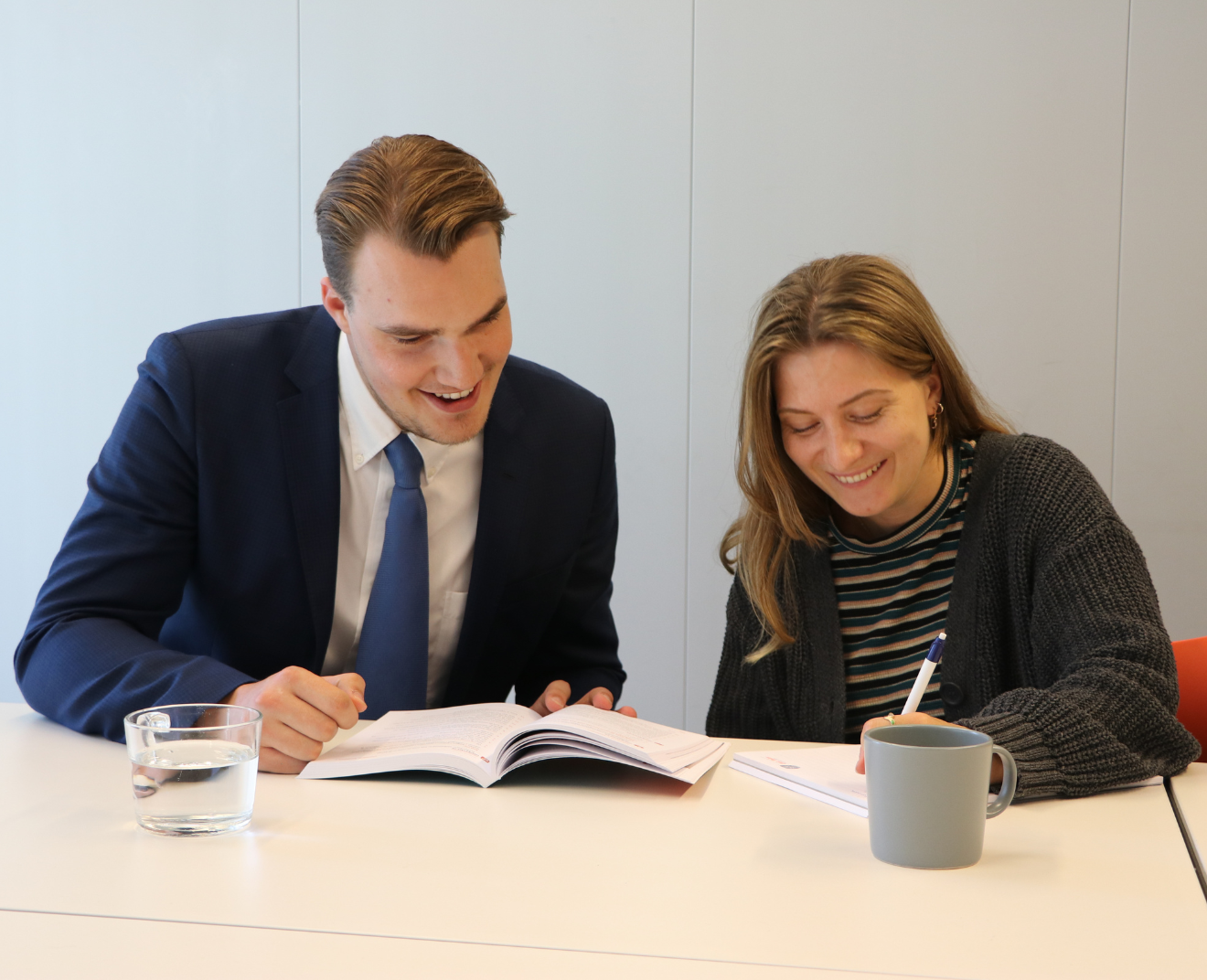 Een man in een donkerblauw pak en een vrouw in een gestreept shirt studeren samen aan een tafel. De man wijst naar een open boek terwijl de vrouw glimlachend luistert en aantekeningen maakt.
