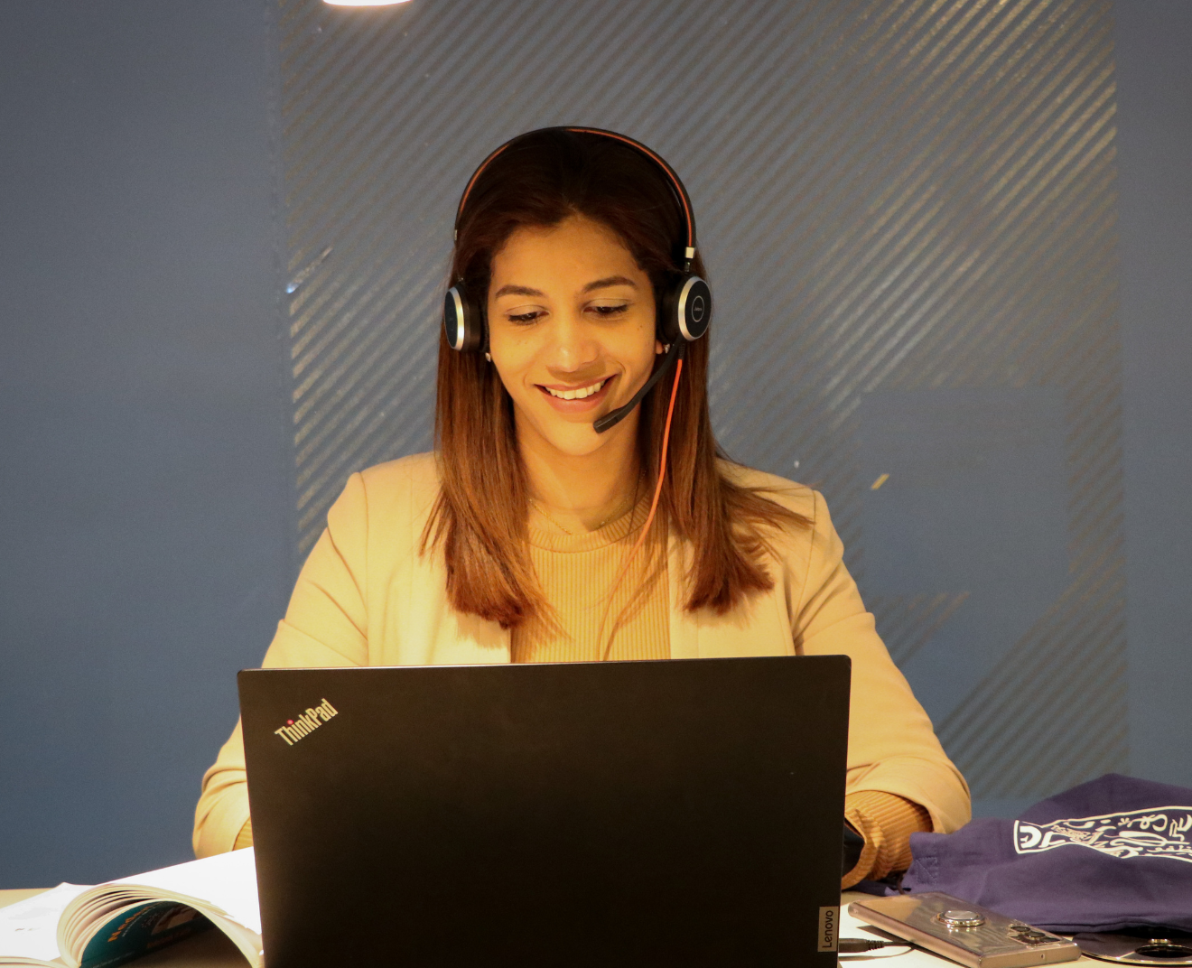 Een lachende vrouw met halflang bruin haar draagt een headset en werkt op een laptop. Ze zit aan een bureau met een open boek en enkele spullen ernaast, in een moderne ruimte met een blauwe wand.