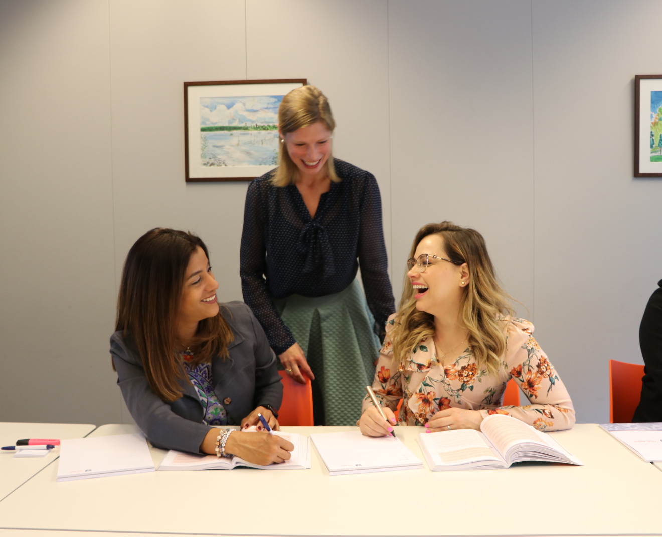 Twee vrouwen zitten aan een tafel met studieboeken en notitieblokken, terwijl een derde vrouw staand met hen meelacht. Ze lijken een plezierig gesprek te hebben.