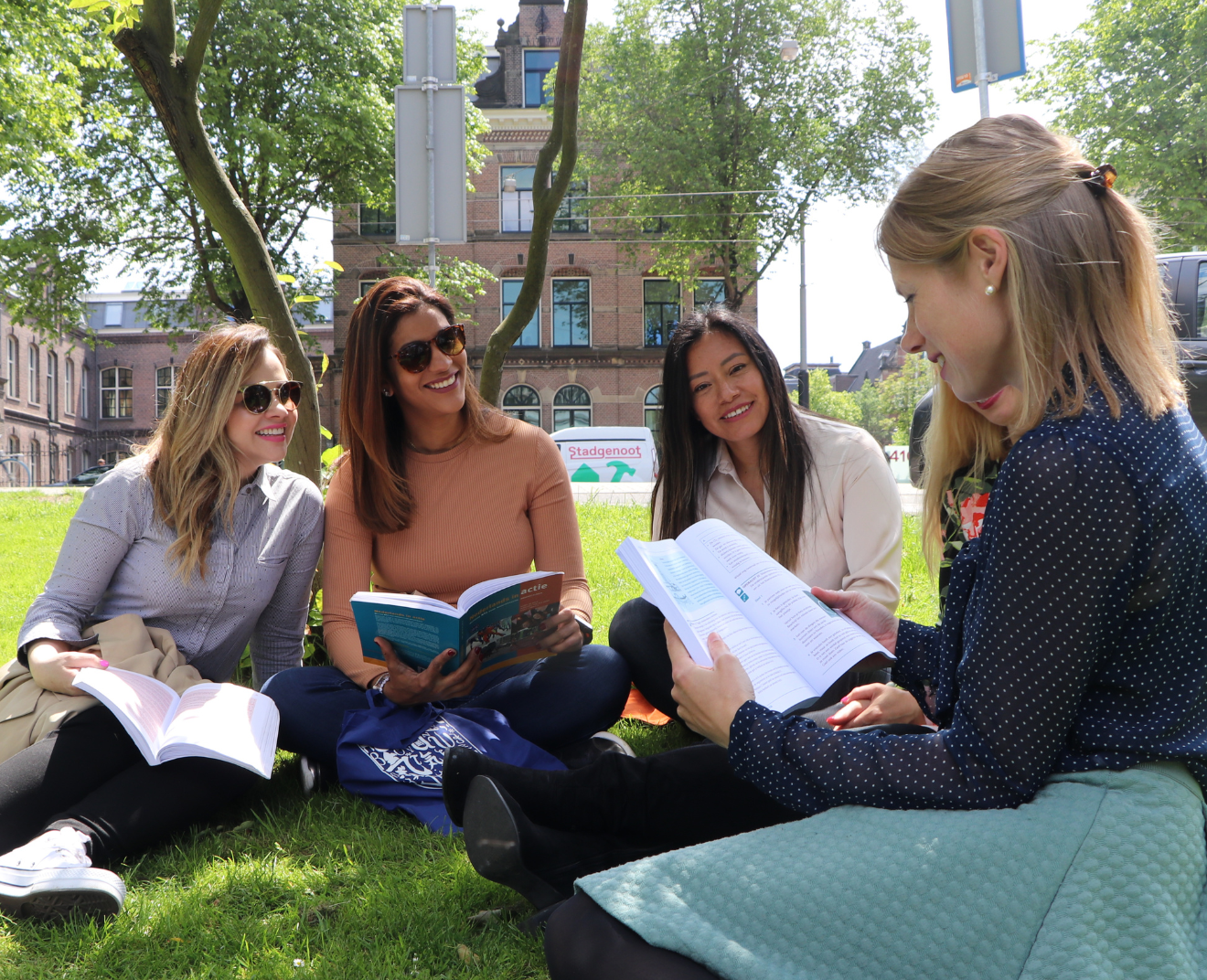 Vier vrouwen zitten op het gras en lezen samen in boeken, lachend en pratend.