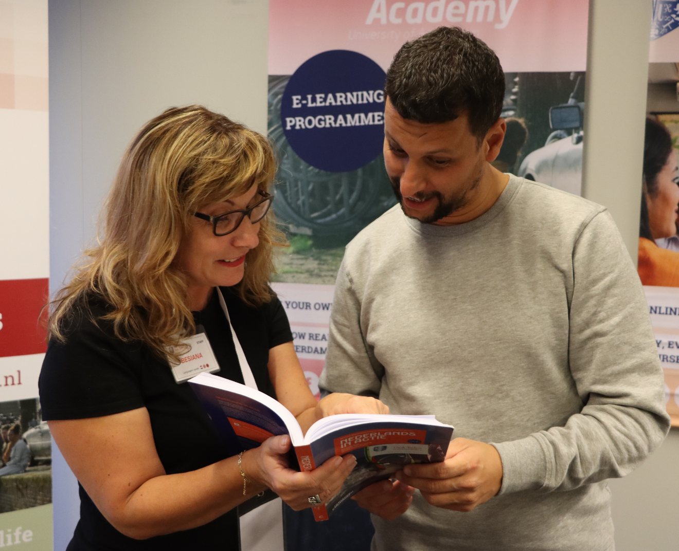 Een vrouw met een naamkaartje en bril laat een man een boek zien, terwijl ze samen lachen. Op de achtergrond staan banners met informatie over e-learningprogramma's.