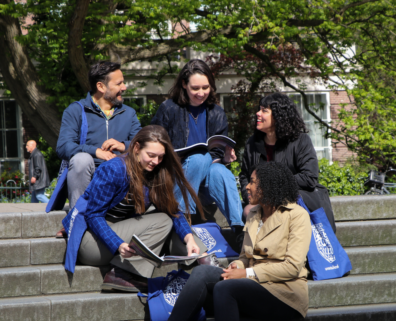 Een groep van vijf mensen zit op een trap, pratend en kijkend in boeken. Ze lachen, met bomen en een bakstenen gebouw op de achtergrond.