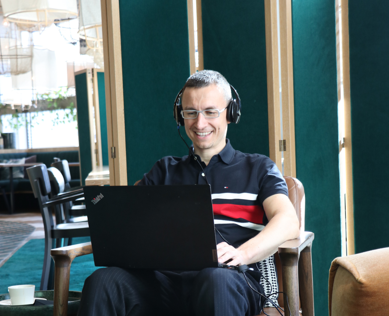 Een lachende man met een bril en headset zit in een comfortabele stoel en werkt op zijn laptop. Hij bevindt zich in een sfeervolle ruimte met groene panelen en houten accenten.