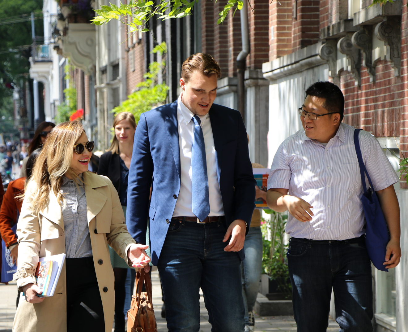 Drie mensen lopen lachend over een zonnige straat met bakstenen gebouwen en groen. De vrouw draagt een beige trenchcoat en houdt boeken vast, terwijl de twee mannen in gesprek zijn en een blauwe tas dragen.