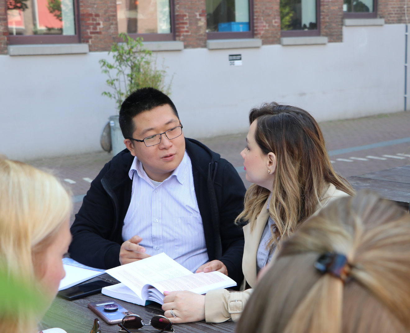 Een man en een vrouw zitten buiten aan een picknicktafel en bespreken een open studieboek. Andere mensen zitten erbij, terwijl de man met bril iets uitlegt en de vrouw aandachtig luistert.