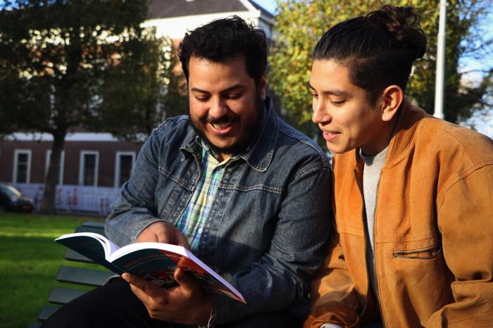 Twee mannen zitten samen buiten op een bankje in een Nederlands studieboek te lezen