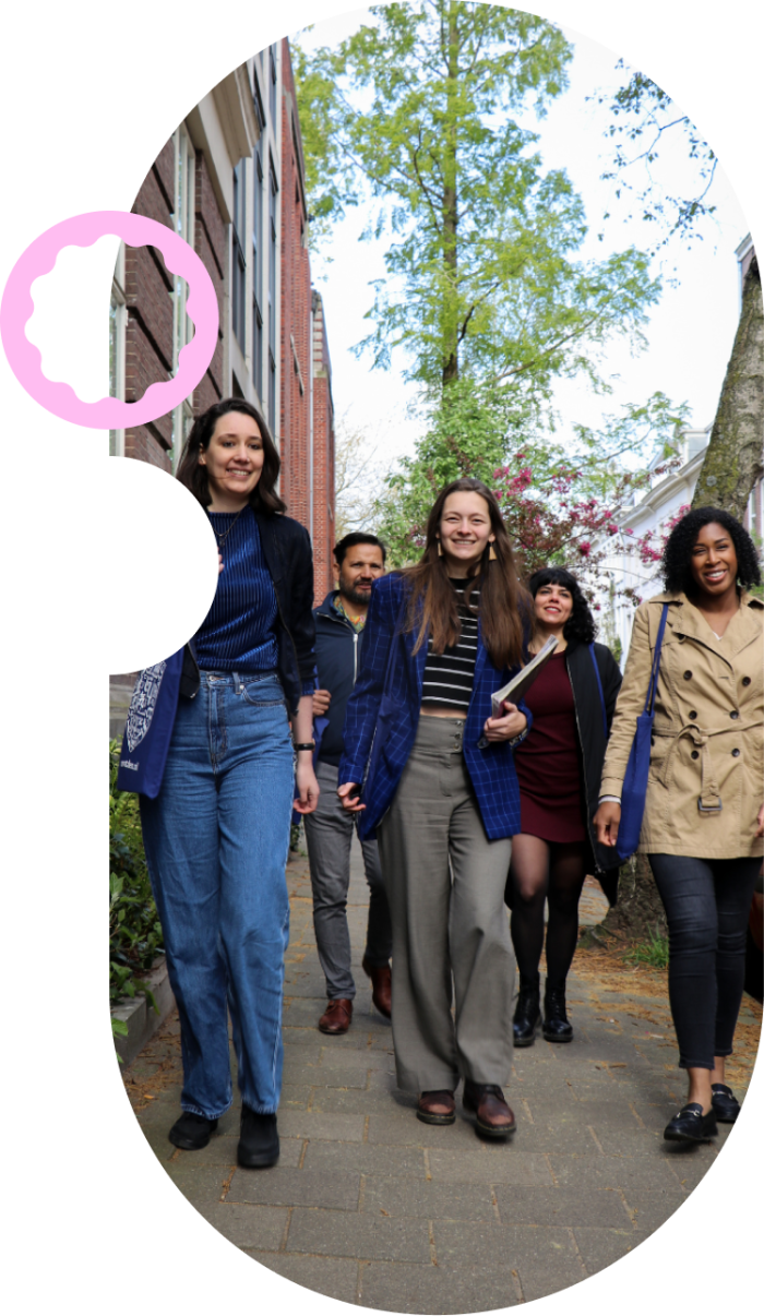 Een groep lachende mensen loopt samen over een straat met bomen en historische gebouwen. Ze dragen tassen en boeken, wat suggereert dat ze op weg zijn naar of van een studie- of werkgerelateerde activiteit.