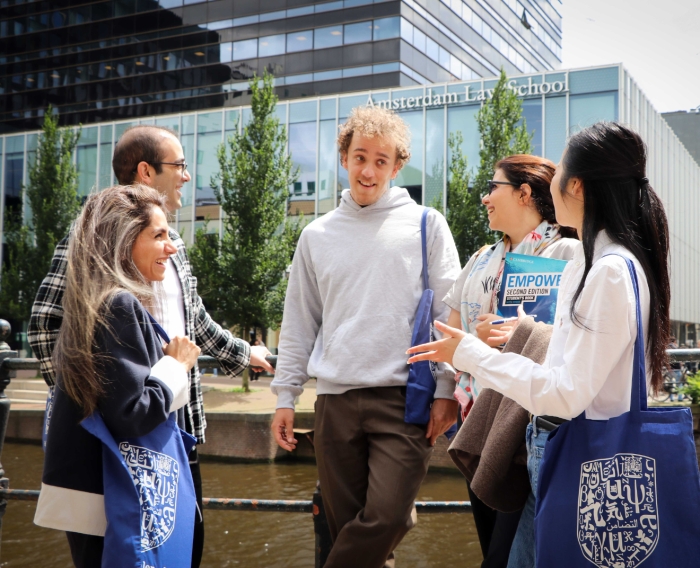 Groep van vijf studenten die buiten bij een gracht staan te praten en lachen, voor een modern glazen gebouw met de tekst “Amsterdam Law School”; sommigen dragen boeken en totebags.