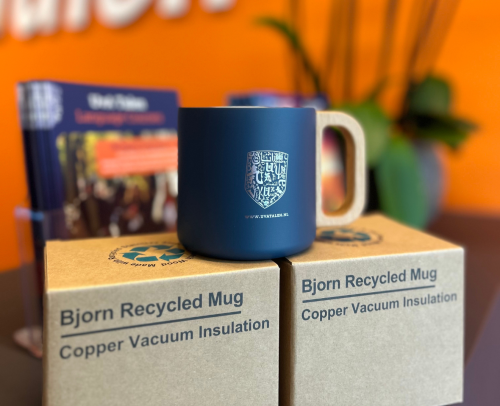 Een donkerblauwe mok met een houten handvat staat op twee gerecyclede kartonnen dozen. In de wazige achtergrond zijn brochures en groene planten te zien tegen een oranje muur.