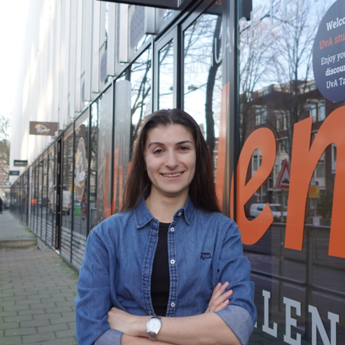 Woman with long brown hair and a denim shirt stands smiling with arms crossed in front of a glass facade with large orange letters. 