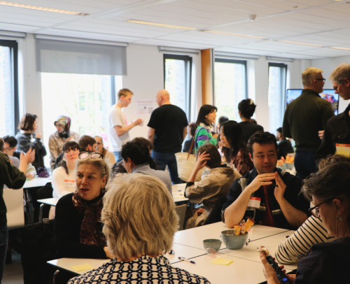 A crowded room with people sitting in groups at tables, actively engaged in conversation. In the background, people are standing and walking around; the atmosphere is lively and social.